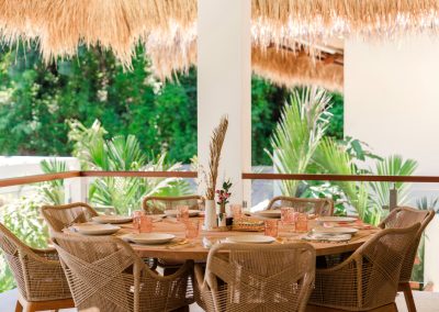 Thatched terrace dining at Villa Svara at Vesica Villas Resort in Amed, with a round wood table, woven rattan chairs, pink-tinted glassware, and lush tropical foliage beyond a glass railing