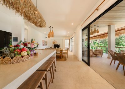 Open living level at Villa Svara at Vesica Villas Resort in Amed, with kitchen island and fringe pendant, wood dining under woven lights, and black-framed doors open to a thatched terrace
