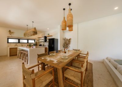 Indoor dining at Villa Svara with a long wood table, six woven-seat chairs on a jute rug, three bottle-shaped wicker pendants, kitchen island to the left, and marble-tiled floors