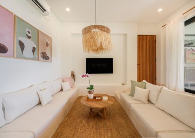 Sunken media lounge at Villa Svara with facing built-in sofas, pink and sage pillows, round wood coffee table, large straw-fringe pendant, framed Balinese art, and a wall-mounted TV