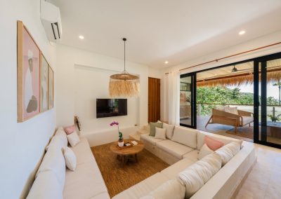Sunken lounge at Villa Svara at Vesica Villas Resort in Amed, with cream cushions and pink green pillows, straw pendant, wall art and TV, open to a thatched balcony overlooking tropical hillside and sea