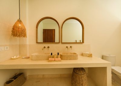 Double vanity close-up at Villa Svara with fluted stone vessel sinks, arched wood mirrors, brass faucets, pink towels on a tray, woven fringe pendant, and marble floor