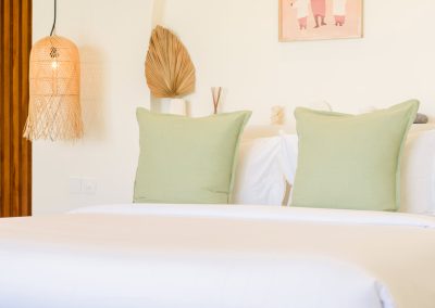 King bed headboard detail at Villa Svara with sage green pillows, woven pendant lamp, shelf with dried palm fan and diffuser, and soft pink-toned framed art above