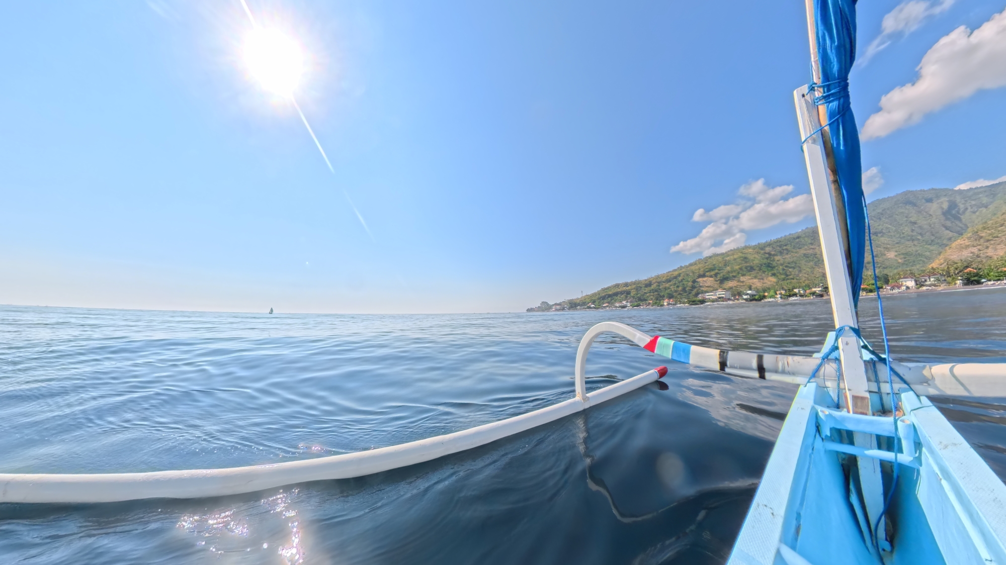 Bright sun over calm blue water from a jukung outrigger boat off Amed, with green hills and villages along the East Bali coast.