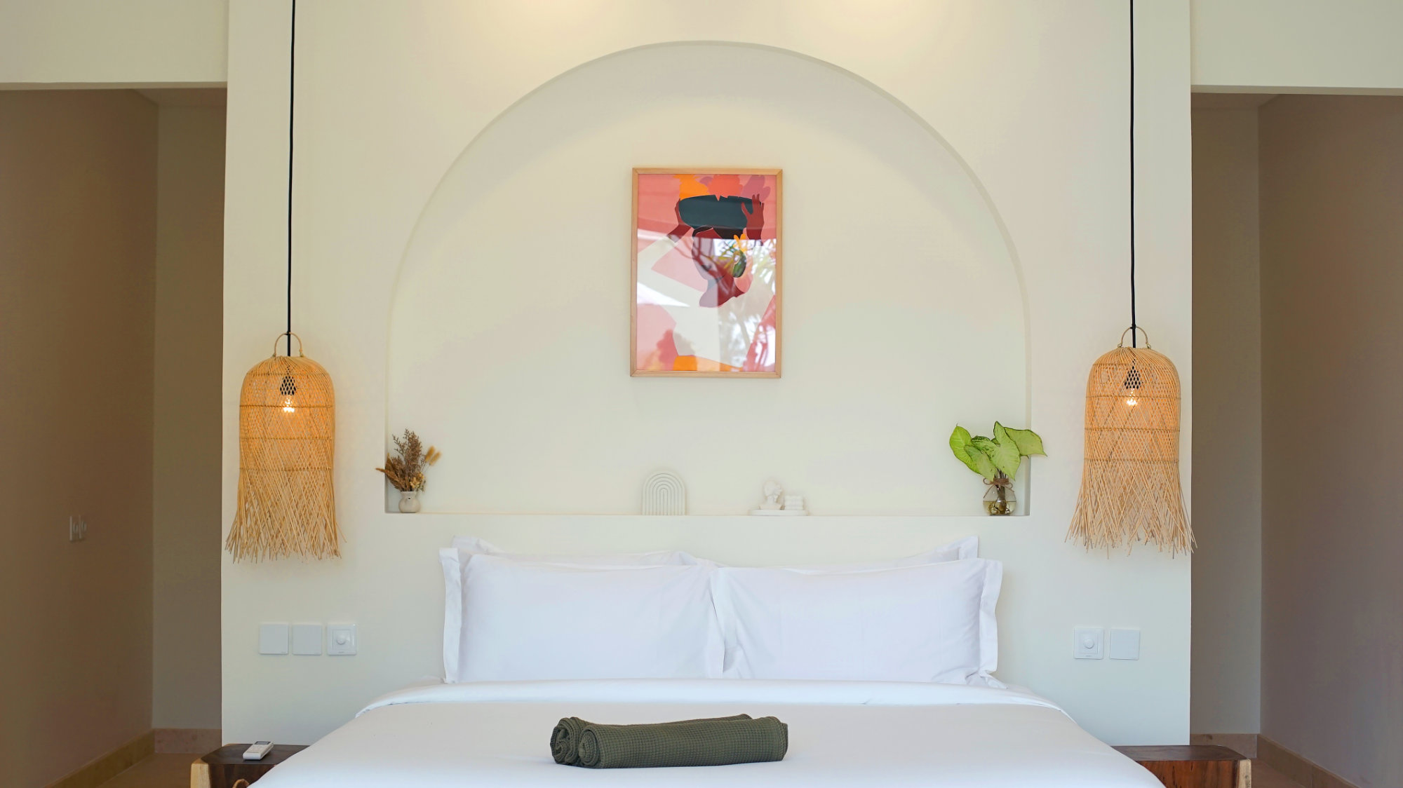 White king bed beneath an arched alcove with colourful artwork, rattan pendant lights and a folded green towel, in Villa Svara at Vesica Villas Resort, Amed.